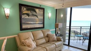 Un salon avec un canapé beige, un tableau encadré représentant une scène de plage et une vue sur l’océan par une porte-fenêtre coulissante.