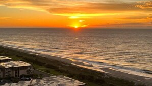 Un coucher de soleil sur l’océan, avec une plage et un bâtiment visibles au premier plan.