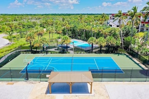 A tennis court with a net, surrounded by a fence, and a covered seating area in the foreground.