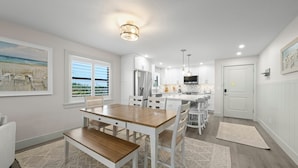 A modern kitchen with white cabinetry, a central island, and a dining area with a wooden table and chairs. - Tarpon Beach 206: Beautiful Condo & Stunning Gulf Views to Match (Sanibel)