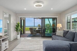 A modern living room with a grey sofa, a flat-screen TV, a dining area with a table and chairs, and a balcony with a view of the ocean.