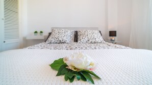 Una cama con cubrecolcha blanco texturizado, dos almohadas con diseño blanco y negro, y una flor blanca con hojas verdes sobre la cama.