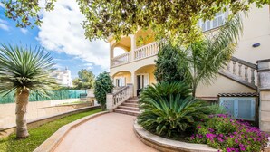 Un edificio de dos pisos con una escalera central, rodeado por un jardín ajardinado con palmeras y plantas en flor.