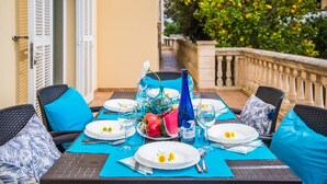 Una mesa de comedor dispuesta con platos, vasos y una botella, rodeada de sillas con cojines azules, en un balcón con barandilla y follaje al fondo.