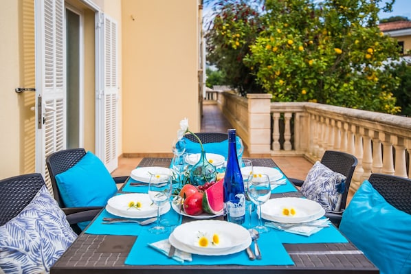 A dining table set with plates, glasses, and a bottle, surrounded by chairs with blue cushions, on a balcony with a railing and greenery in the background.