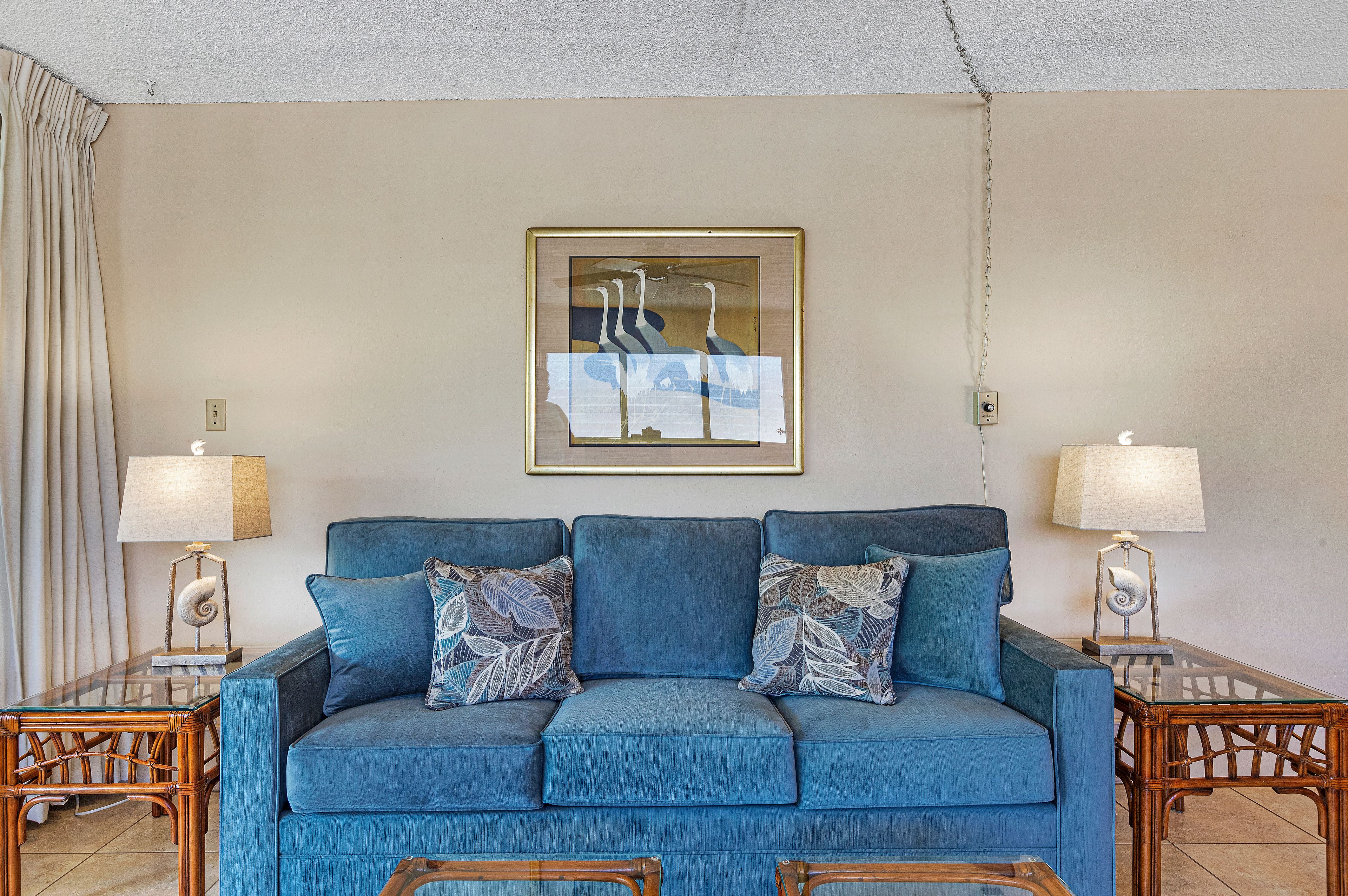 A blue sofa with patterned pillows, two side tables with lamps, a framed artwork above the sofa, and a ceiling light fixture.