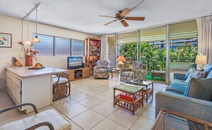 A spacious living area with a kitchen, a dining table, and a sofa. There is a ceiling fan, a bookshelf, and a large window with a view of greenery.