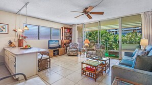 A spacious living area with a kitchen, a dining table, and a sofa. There is a ceiling fan, a bookshelf, and a large window with a view of greenery.
