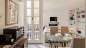 Cuina moderna amb microones i cafetera integrades, taula de vidre per dinar amb cadires, televisió penjada sobre una llar de foc i una prestatgeria amb diversos objectes.