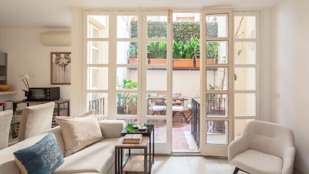 Un saló amb sofà, butaca, taula de centre i vista cap a un pati exterior amb plantes i mobiliari.