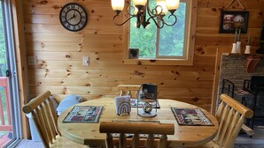 A wooden dining room with a round table, chairs, a chandelier, a clock, and a window with a view of trees. - Cozy Cabin in the Beautiful Upper Michigan (Rock)