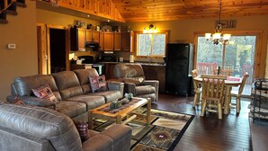 A cozy living room with a leather sofa, a wooden dining table, and a kitchen area with wooden cabinets and a refrigerator. - Cozy Cabin in the Beautiful Upper Michigan (Rock)