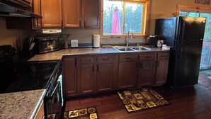 A kitchen with wooden cabinets, a stainless steel sink, a black refrigerator, and a window with a view of trees. - Cozy Cabin in the Beautiful Upper Michigan (Rock)