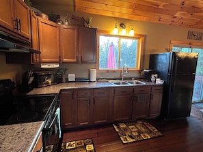 A kitchen with wooden cabinets, a stainless steel sink, a black refrigerator, and a window with a view of trees. - Cozy Cabin in the Beautiful Upper Michigan (Rock)