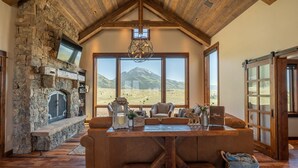 A cozy living room with a stone fireplace, large windows, and a wooden ceiling.