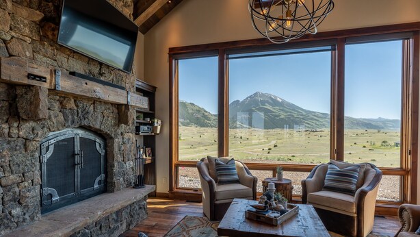 A stone fireplace with a TV above it, two leather armchairs facing a large window with a mountain view.