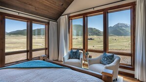 A bedroom with a large bed, two armchairs, and a view of a mountainous landscape through large windows.