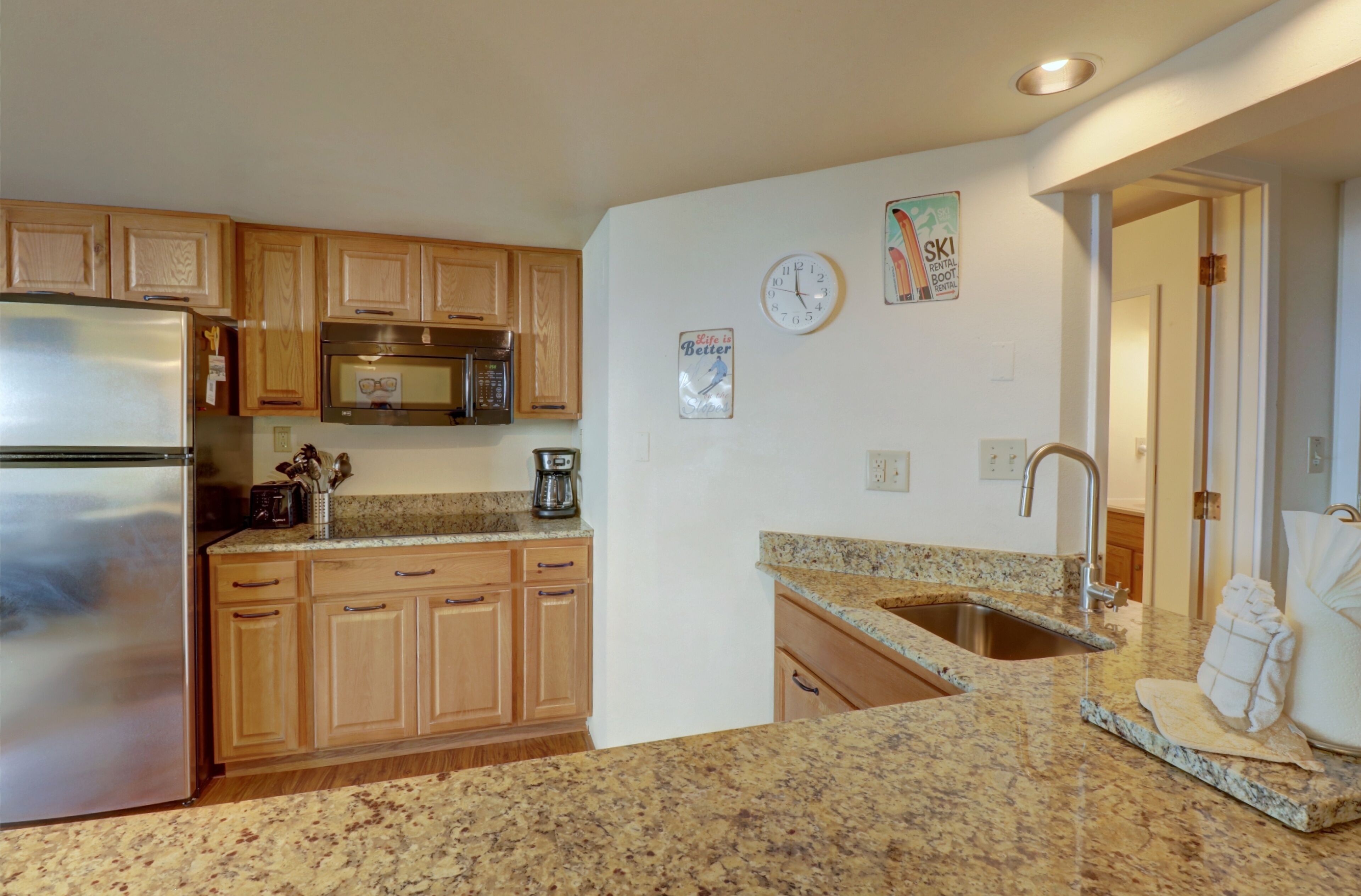 A kitchen with wooden cabinets, a stainless steel refrigerator, granite countertops, a double sink, and a clock on the wall.