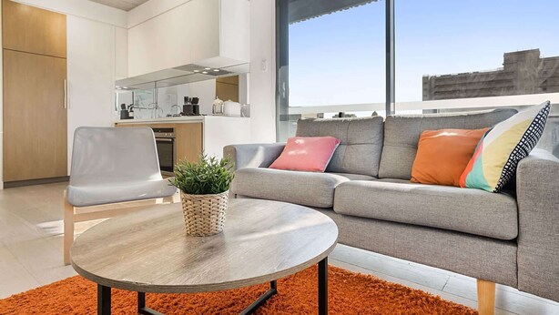 A modern living room with a grey sofa, a round coffee table, and a large window offering a city view. - Comfy 2-Bed Balcony Unit near Iconic Chapel St (Prahran)