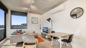 A modern living room with a sofa, chairs, a dining table, a TV, and a large window offering a city view. - Comfy 2-Bed Balcony Unit near Iconic Chapel St (Prahran)