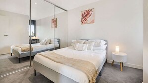 A modern bedroom with a large mirror, a bed with white linens, a bedside table with a lamp, and artwork on the wall. - Comfy 2-Bed Balcony Unit near Iconic Chapel St (Prahran)