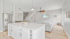 Una cocina moderna con armarios blancos, una isla central, electrodomésticos de acero inoxidable, iluminación colgante y un ventilador de techo.