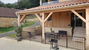 A two-story wooden house with a covered patio, outdoor seating, and a stone wall in the background. - Le Séchoir des acacias, 110m2 gîte "La Noyeraie" with heated swimming pool. (SAINT-LEON-SUR-VEZERE)