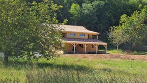 A two-story wooden house with a tiled roof, surrounded by lush greenery and trees. - Le Séchoir des acacias, 110m2 gîte "La Noyeraie" with heated swimming pool. (SAINT-LEON-SUR-VEZERE)
