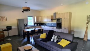 A compact living space with a grey sofa, yellow cushions, a dining table, and a kitchen area with wooden cabinets and appliances. - Le Séchoir des acacias, 110m2 gîte "La Noyeraie" with heated swimming pool. (SAINT-LEON-SUR-VEZERE)