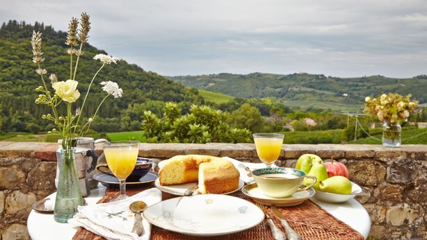 A table set with breakfast items, including a muffin, a glass of orange juice, and a plate with a pear and apple, overlooking a scenic landscape. - [VILLA ACQUAPIETRA FLORENCE⭐⭐⭐⭐⭐] Pool | Vineyards | Olive trees (San Casciano Val di Pesa)