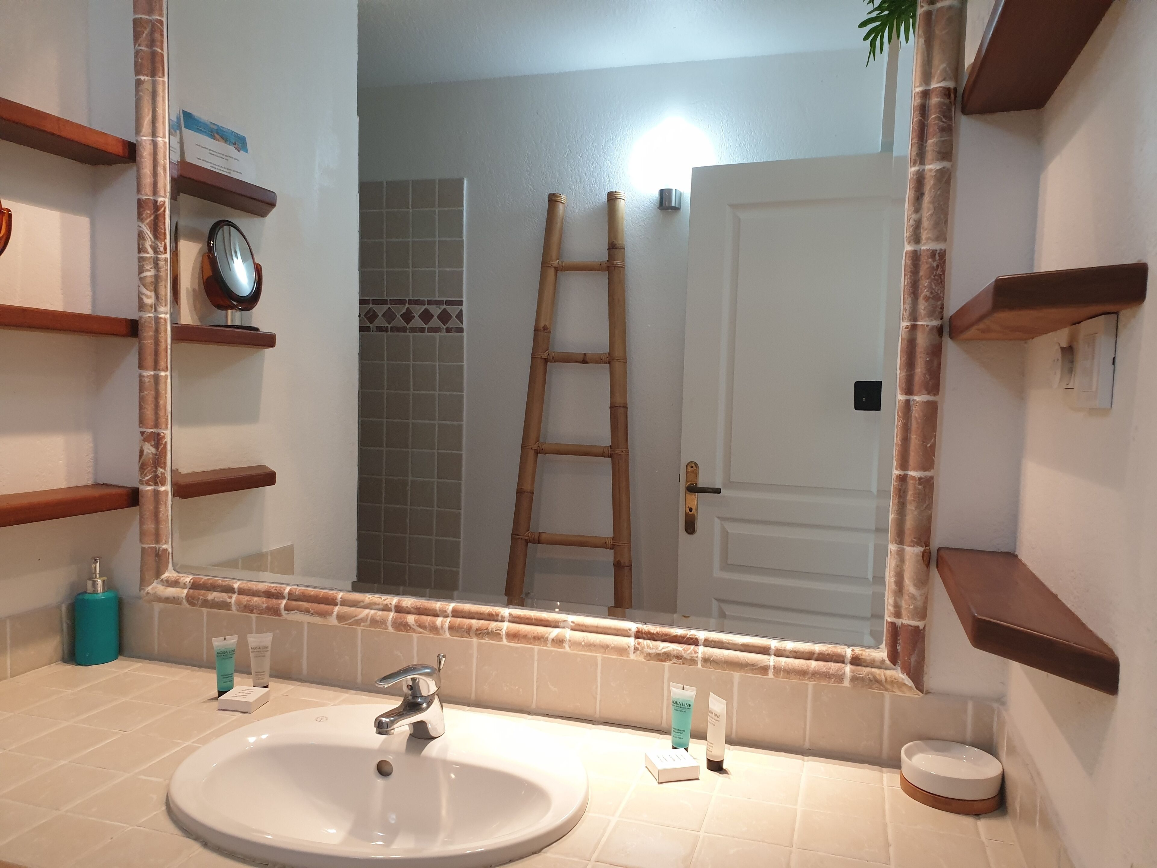 Un baño con un espejo grande, un lavamanos, artículos de aseo y una escalera de madera apoyada en la pared.