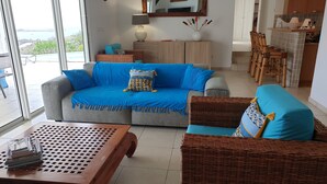 A living room with a grey sofa, a blue sofa, and a wicker chair. There is a wooden table with magazines, a lamp, and a vase with dried flowers. A large window offers a view of the outdoors. - Villa JELUCA Beautiful and spacious villa with swimming pool (SAINT MARTIN)