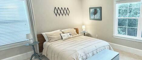 A bedroom with a bed, bedside table, lamp, artwork, and a window.