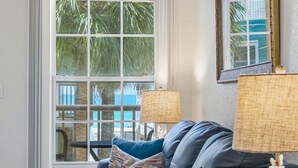 A living room with a sofa, a lamp, a small table, and a large window with a view of palm trees.