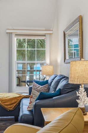 A living room with a sofa, a lamp, a small table, and a large window with a view of palm trees.