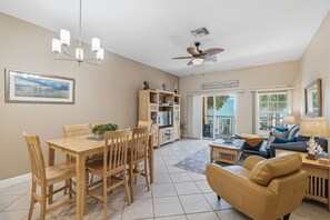 A spacious living area with a dining table, wooden chairs, a sofa, a ceiling fan, and a bookshelf.