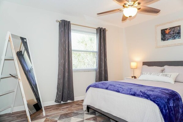 Un dormitorio con cama, ventilador de techo, ventana con cortinas, mesita de noche con lámpara y un cuadro en la pared.