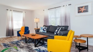 A modern living room with a sofa, coffee table, and chairs. There are windows with curtains, a lamp, and a framed picture on the wall. - New Modern Chic Suite Walkable Steps to Beach (Indian Rocks Beach)