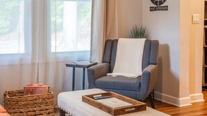 A cozy room with a grey armchair, a small table with a tray, a wicker basket, and a wall clock.