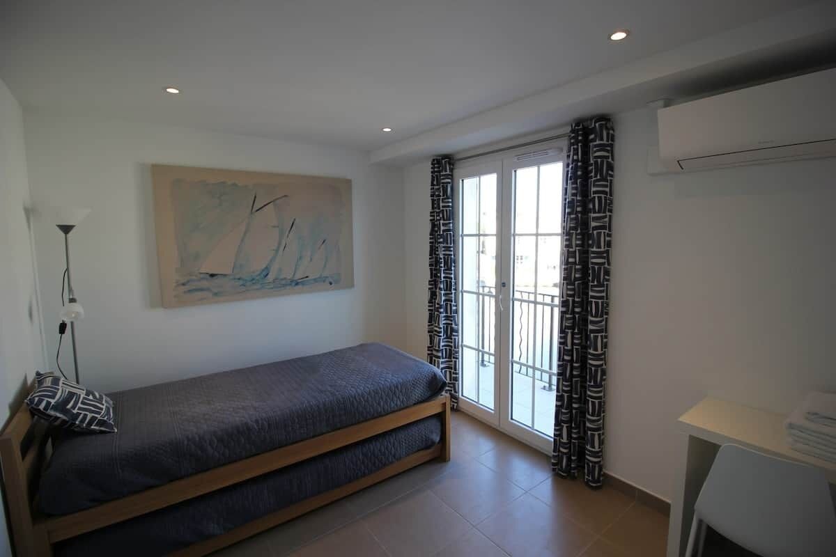 Ein Schlafzimmer mit einem Bett, einem Gemälde, einem Fenster mit gemusterten Vorhängen und einem Tisch mit einer Lampe.