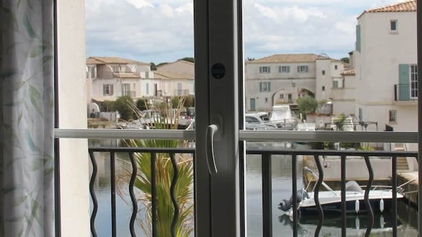 Une vue à travers une fenêtre avec un balcon, une rambarde, une porte et une vue sur une marina avec des bateaux et des bâtiments.
