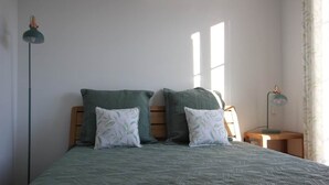 Una cama con colcha estampada, dos almohadas, una mesita de noche, una lámpara y una ventana con cortinas.