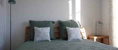 Een bed met een geborduurd sprei, twee kussens, een nachtkastje, een lamp en een raam met gordijnen.
