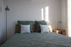 Ein Bett mit einem gemusterten Überwurf, zwei Kissen, ein Nachttisch, eine Lampe und ein Fenster mit Vorhängen.