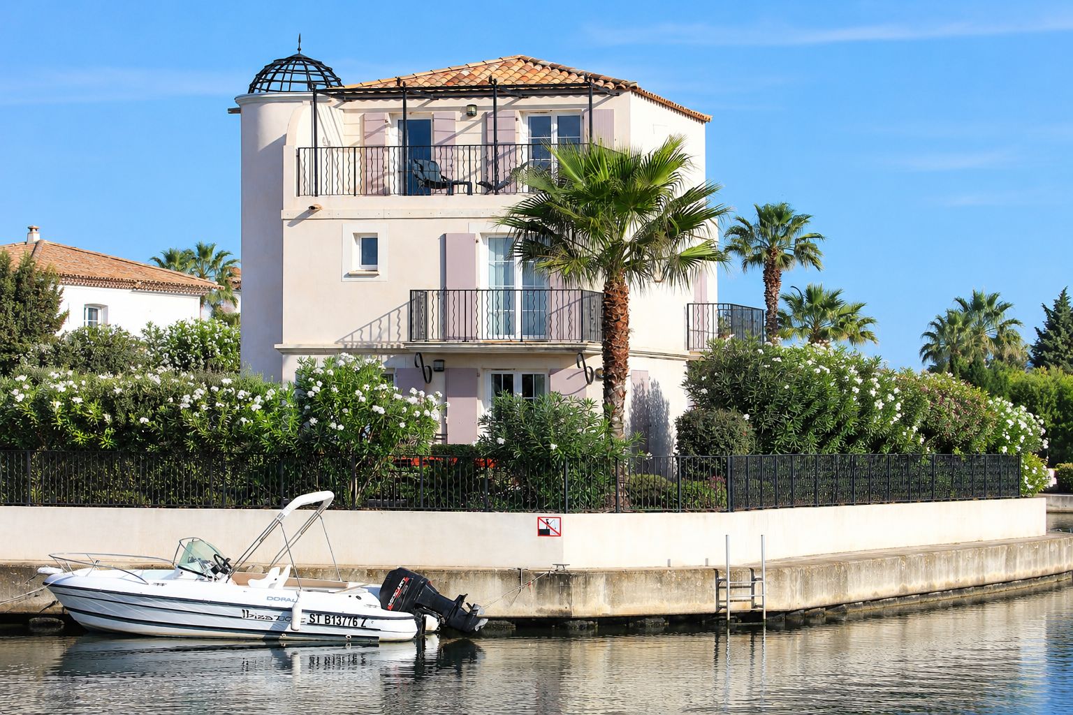 A two-story building with balconies, a boat docked in front, and a palm tree.