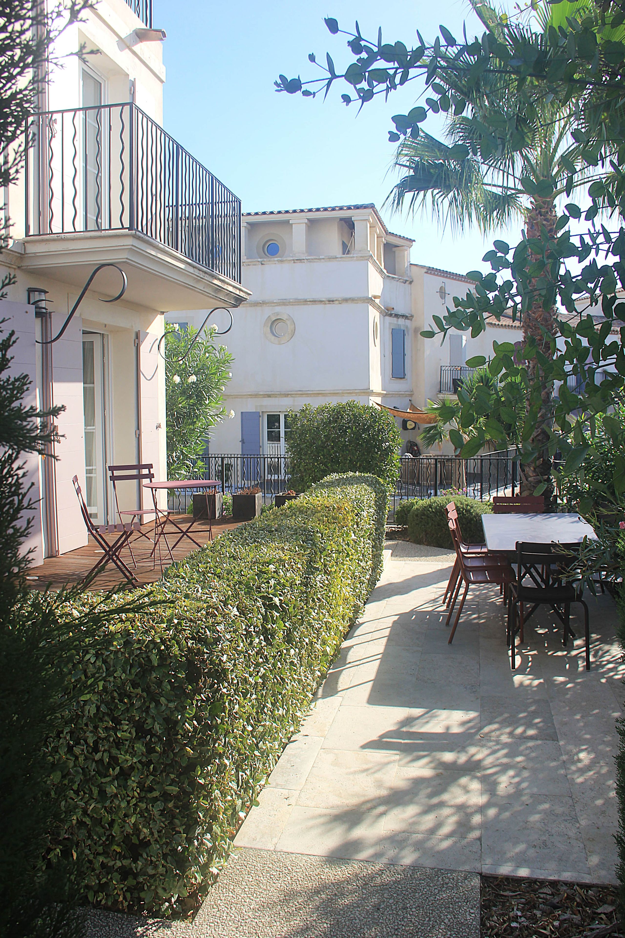 A courtyard with a table and chairs, a neatly trimmed hedge, and a building with a balcony.