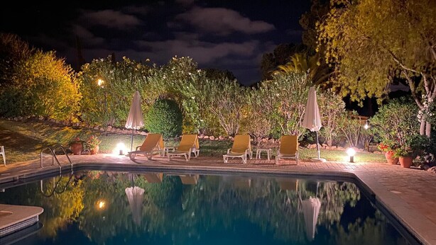 Ein Poolbereich bei Nacht mit Liegestühlen und Sonnenschirmen, umgeben von Bäumen und Pflanzen.