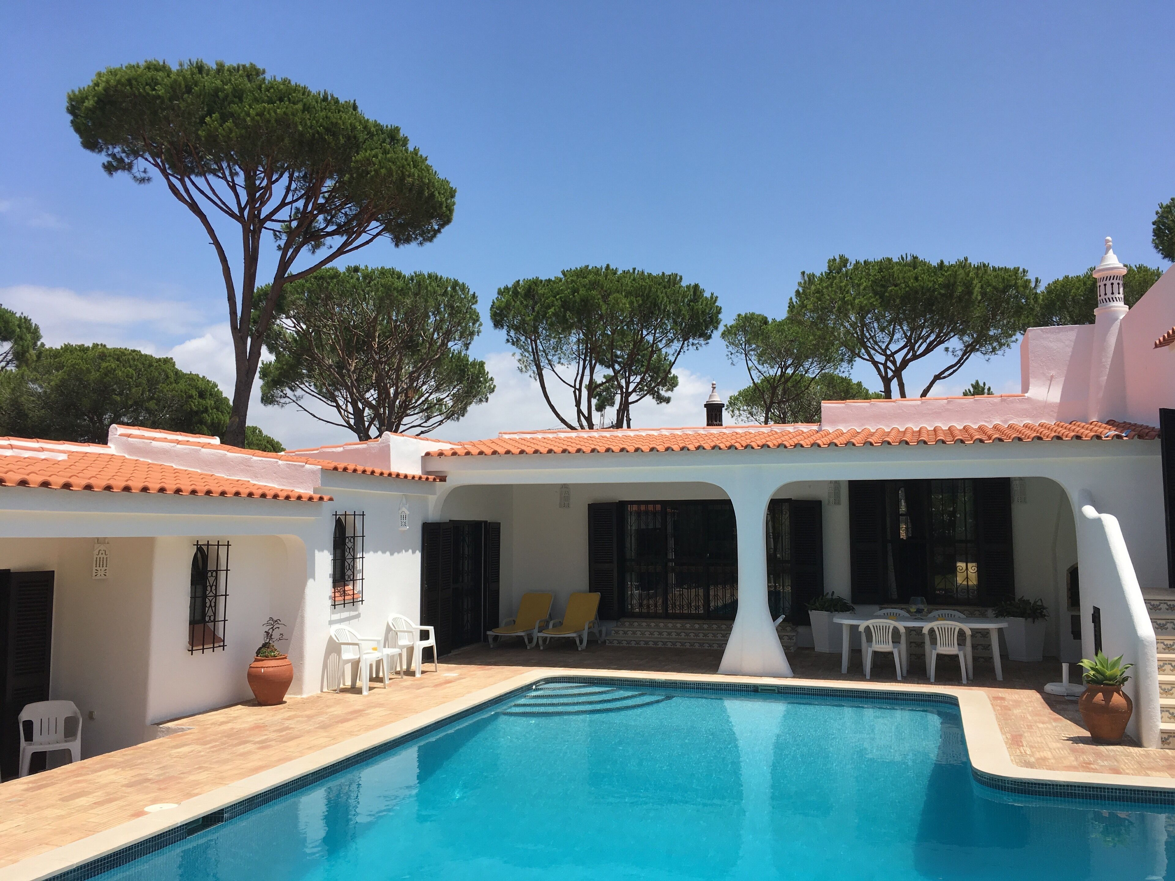Un espace piscine avec une piscine à l’eau d’un bleu limpide, des murs blancs et un toit recouvert de tuiles. On y trouve des chaises et une table d’extérieur.