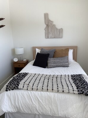Un lit bien fait, avec une couette blanche, un plaid à motifs et plusieurs oreillers. On y trouve une tête de lit en bois, une table de chevet avec une lampe et une œuvre d’art abstraite accrochée au mur.