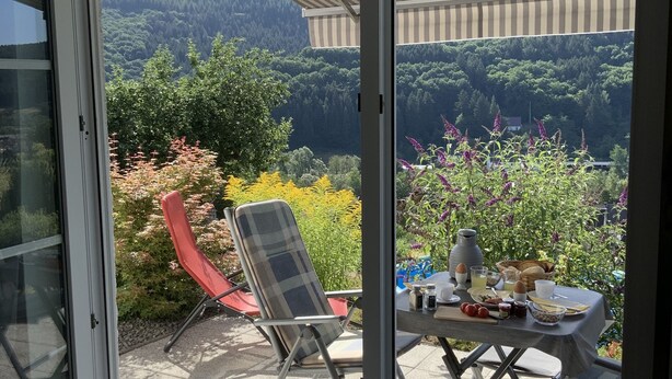 Une terrasse avec une table préparée pour le petit-déjeuner, une chaise et une vue sur une colline boisée.